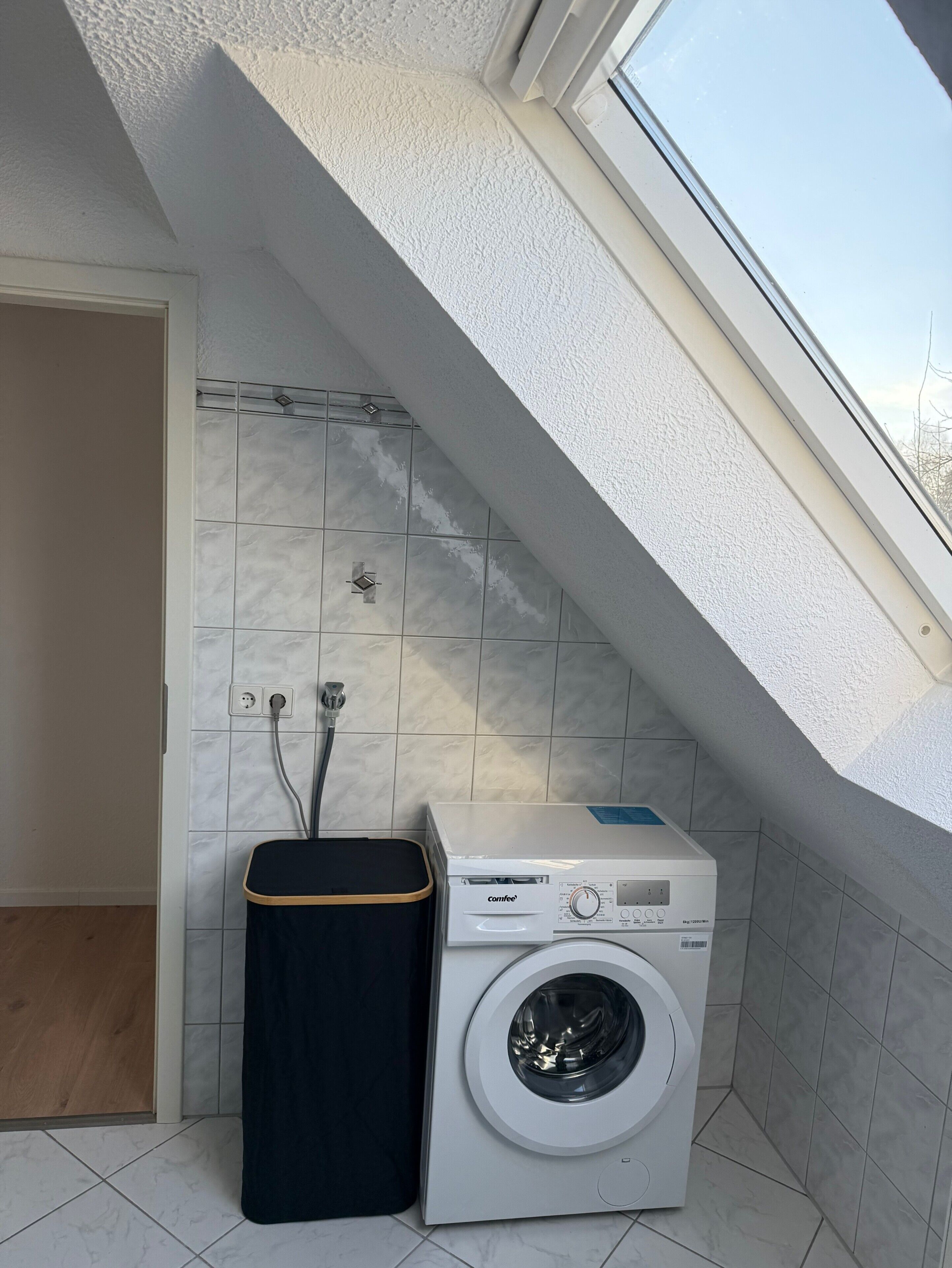 Un lavadero pequeño con lavadora, un cubo de basura y un muro cubierto de azulejos.