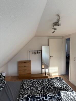 Une chambre avec un plafond en pente, un lit recouvert d’une couvre-lit à motifs zébrés, une commode en bois, un miroir et un luminaire fixé au plafond.