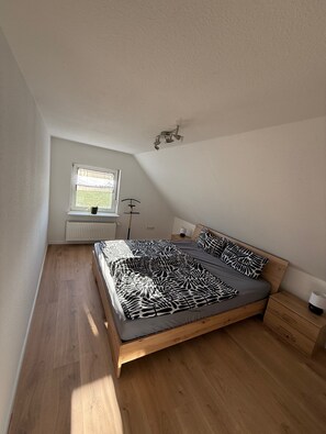 A bedroom with a sloped ceiling, a window with a radiator, a bed with a patterned bedspread, and a bedside table with a lamp. - Apartment 'Waldblick Alterkülz' with Wi-Fi (Alterkülz)