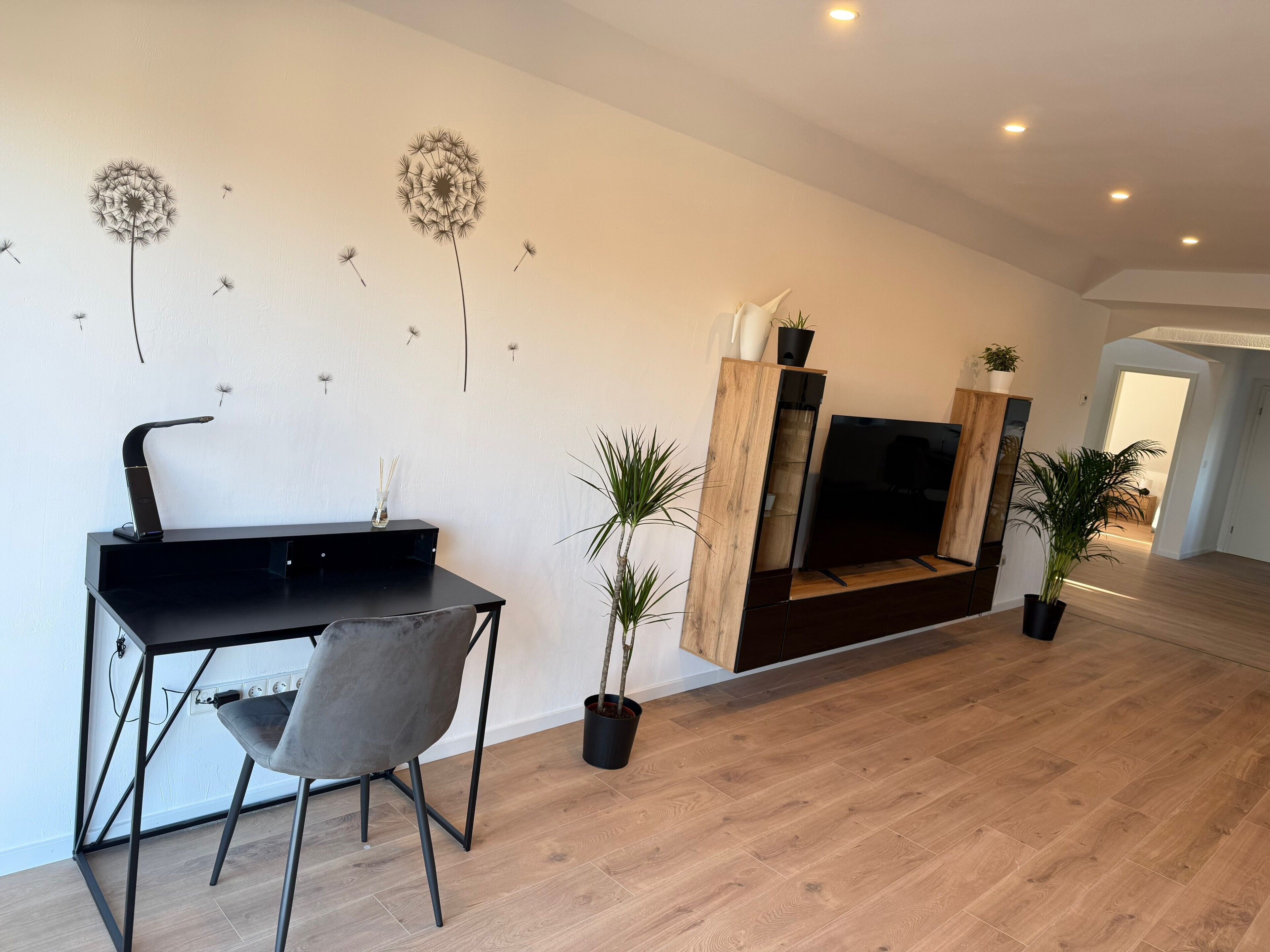 A modern living room with a black desk, grey chair, wooden floor, TV stand, and potted plants.