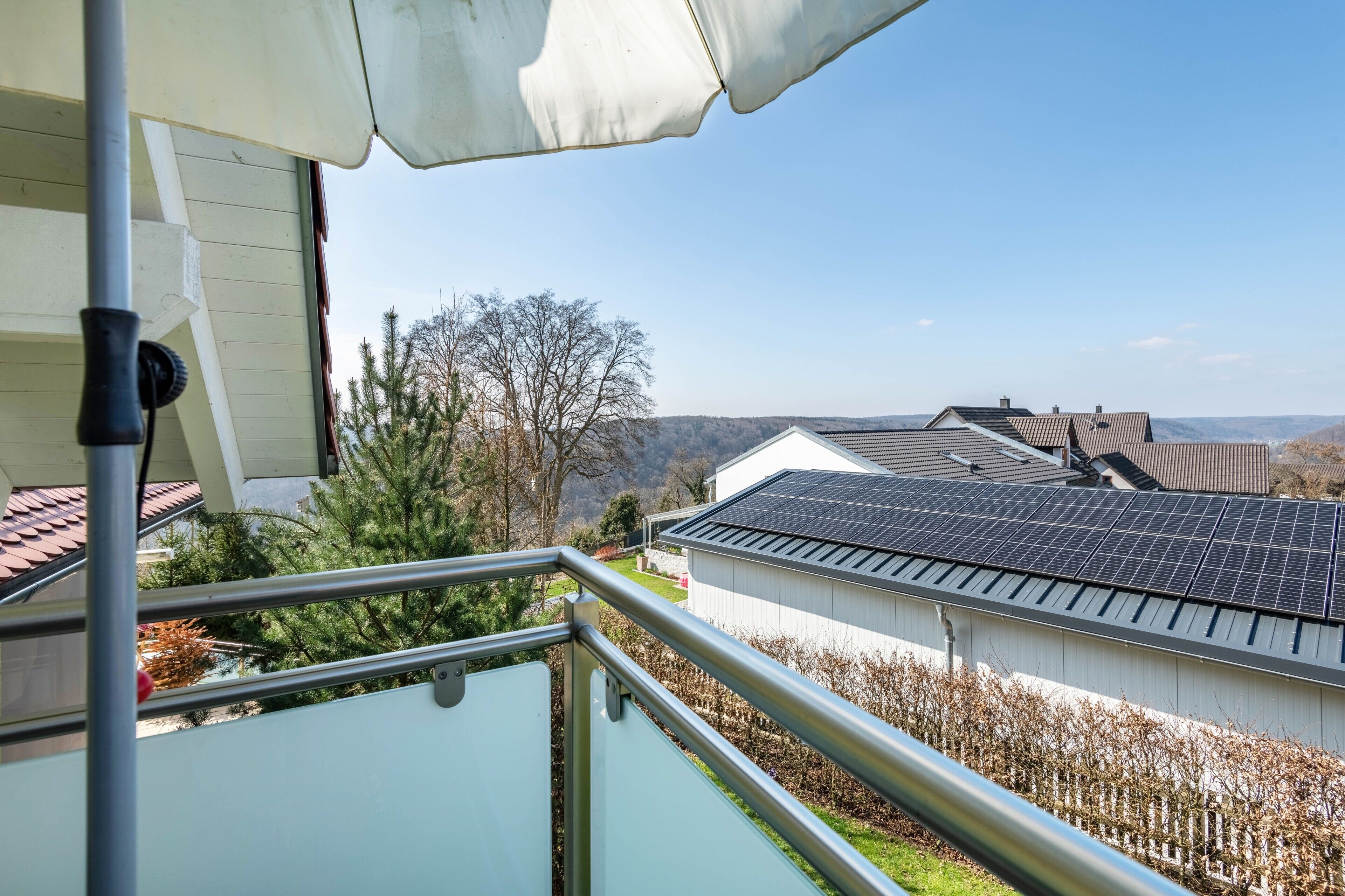 Un balcón con barandilla metálica, una sombrilla blanca y vistas a paneles solares en edificios cercanos.