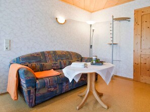 A room with a patterned sofa, a small table with a white tablecloth, a floor lamp, and a wooden door. - Holiday Apartment 'for 2 People' with Shared Garden, Balcony and Wi-Fi (Bad Rippoldsau-Schapbach)