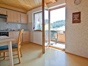 A kitchen with wooden cabinets, a dining table with chairs, a window with a view of trees, and a clock on the wall. - Holiday Apartment 'for 2 People' with Shared Garden, Balcony and Wi-Fi (Bad Rippoldsau-Schapbach)