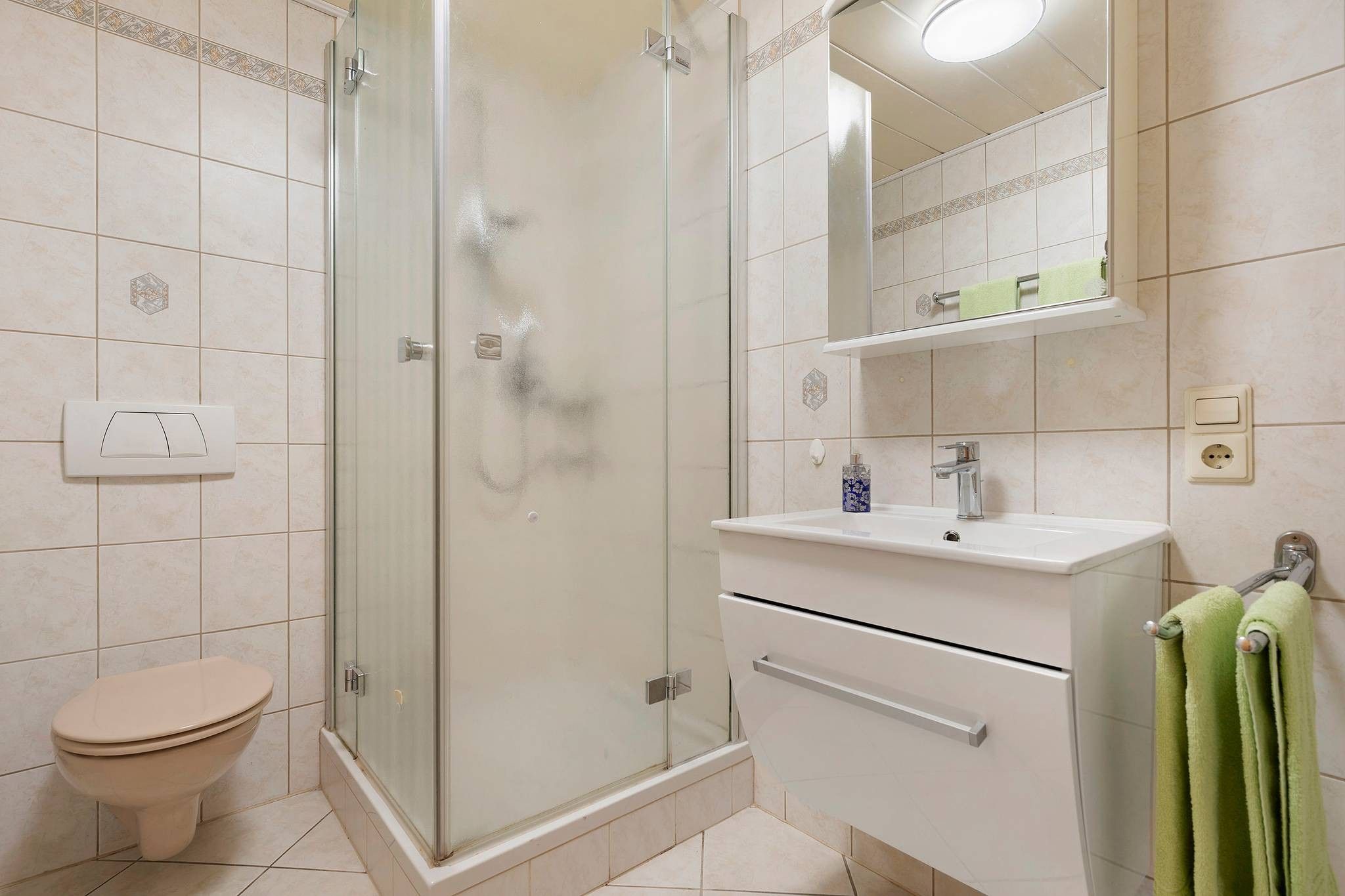 Ein Badezimmer mit einer Duschkabine, Toilette, Waschbecken mit Spiegel darüber und einem Handtuchhalter mit grünen Handtüchern.