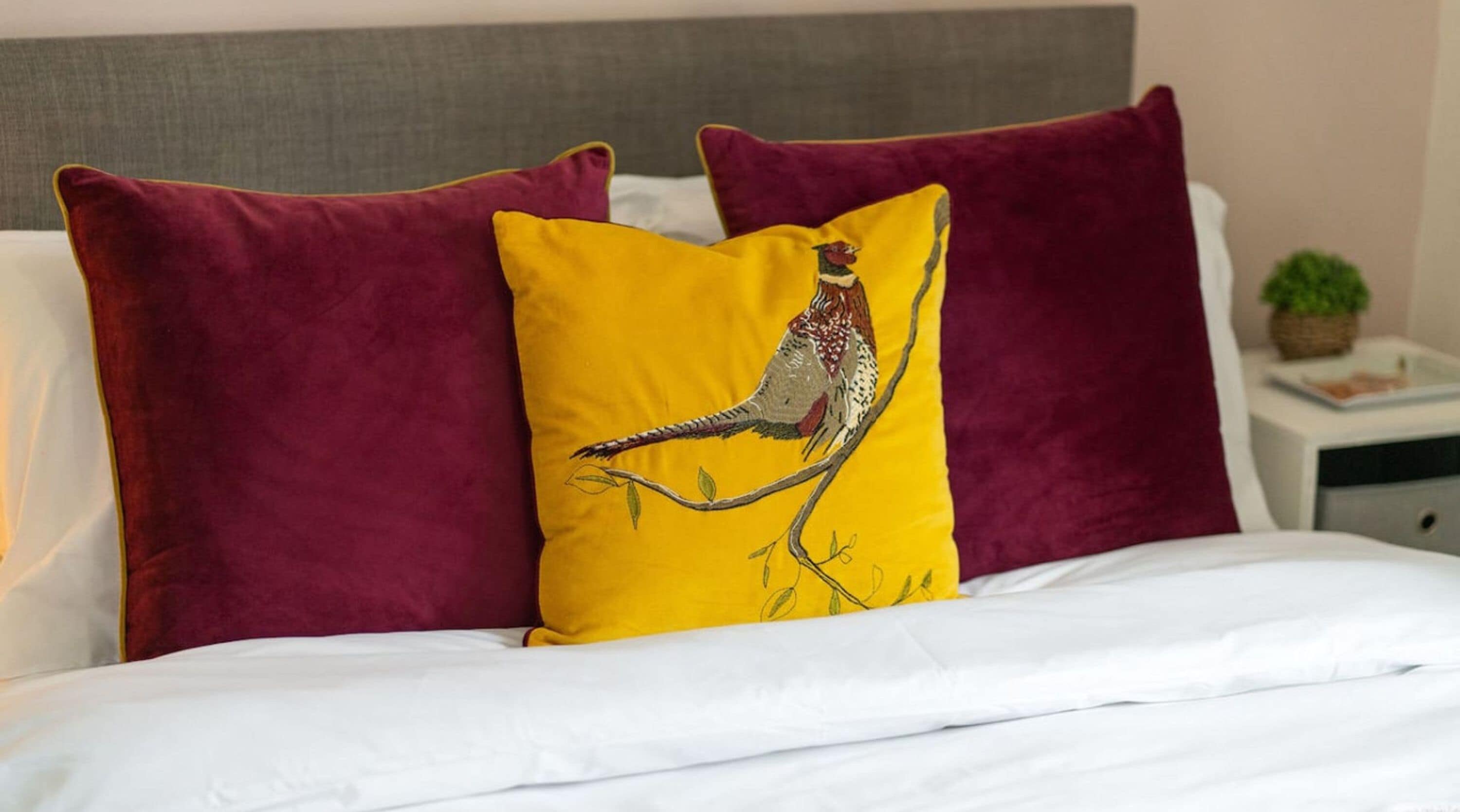 A bed with white bedding, two maroon pillows, and a yellow pillow with a bird design.