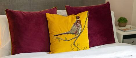 A bed with white bedding, two maroon pillows, and a yellow pillow with a bird design.