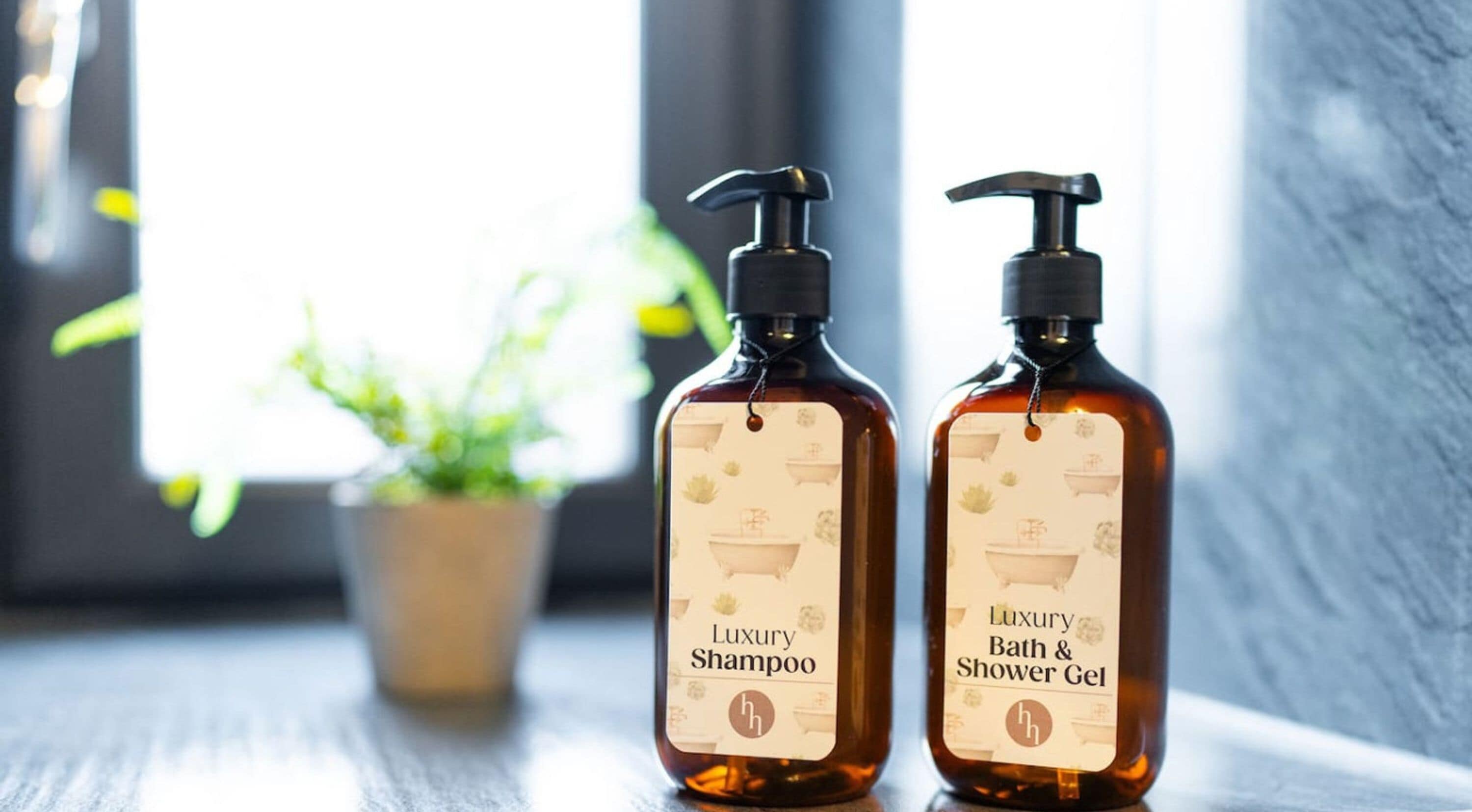 Two bottles of luxury shampoo and bath & shower gel on a wooden surface near a window with a plant in the background.