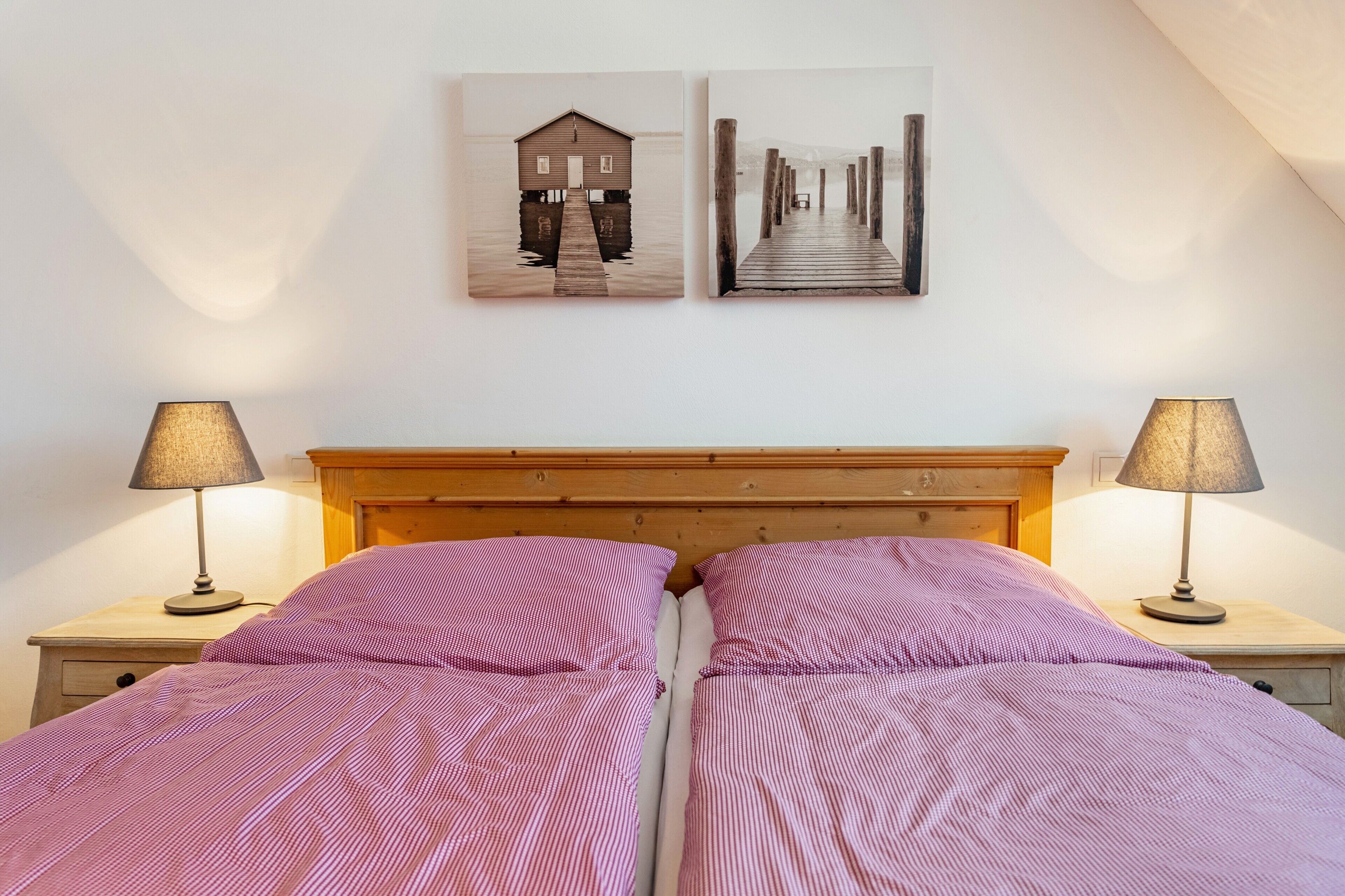 Een bed met een houten hoofdbord, twee nachtkastjes met lampen, en twee kunstwerken aan de muur die een huis en een pier voorstellen.