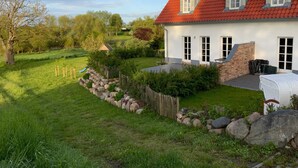 Une maison avec un toit rouge, des murs extérieurs blancs et des fondations en pierre. On y trouve un jardin avec une clôture en bois, un petit étang et une terrasse en bois.