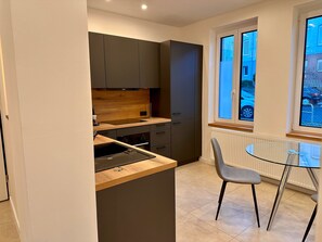 Une cuisine de taille réduite avec des meubles de couleur sombre, un plan de travail en bois et une table en verre accompagnée d’une chaise.