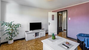 Ein Wohnzimmer mit Fernseher, weißen Möbeln, einer Zimmerpflanze und einer Tür zu einem anderen Raum.