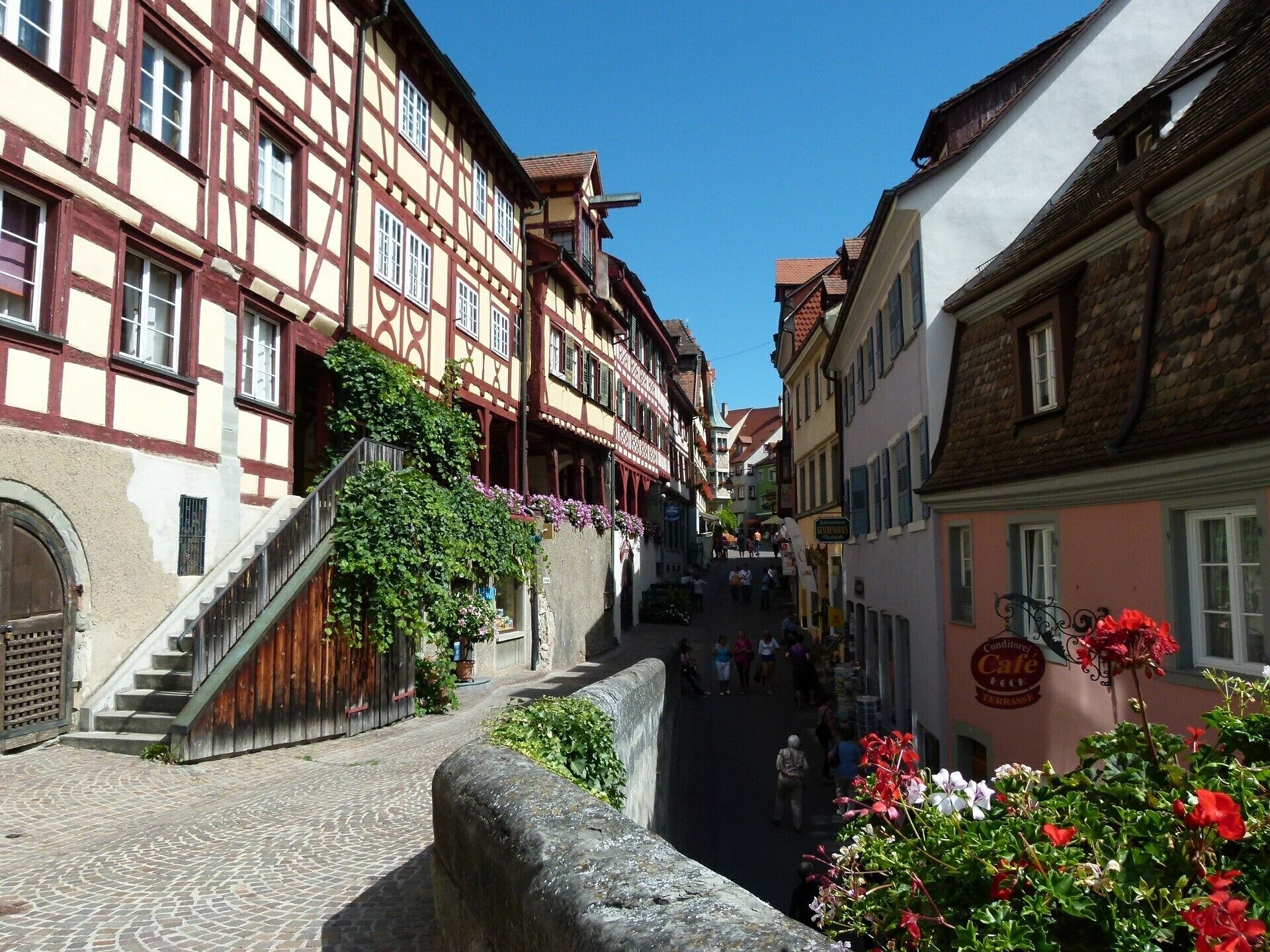 A narrow cobblestone street flanked by traditional half-timbered buildings, a stone wall, and a café sign.