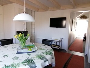 Un comedor con una mesa redonda, sillas, mantel blanco, un jarrón con flores, una lámpara colgante, un televisor en la pared y una alfombra roja.