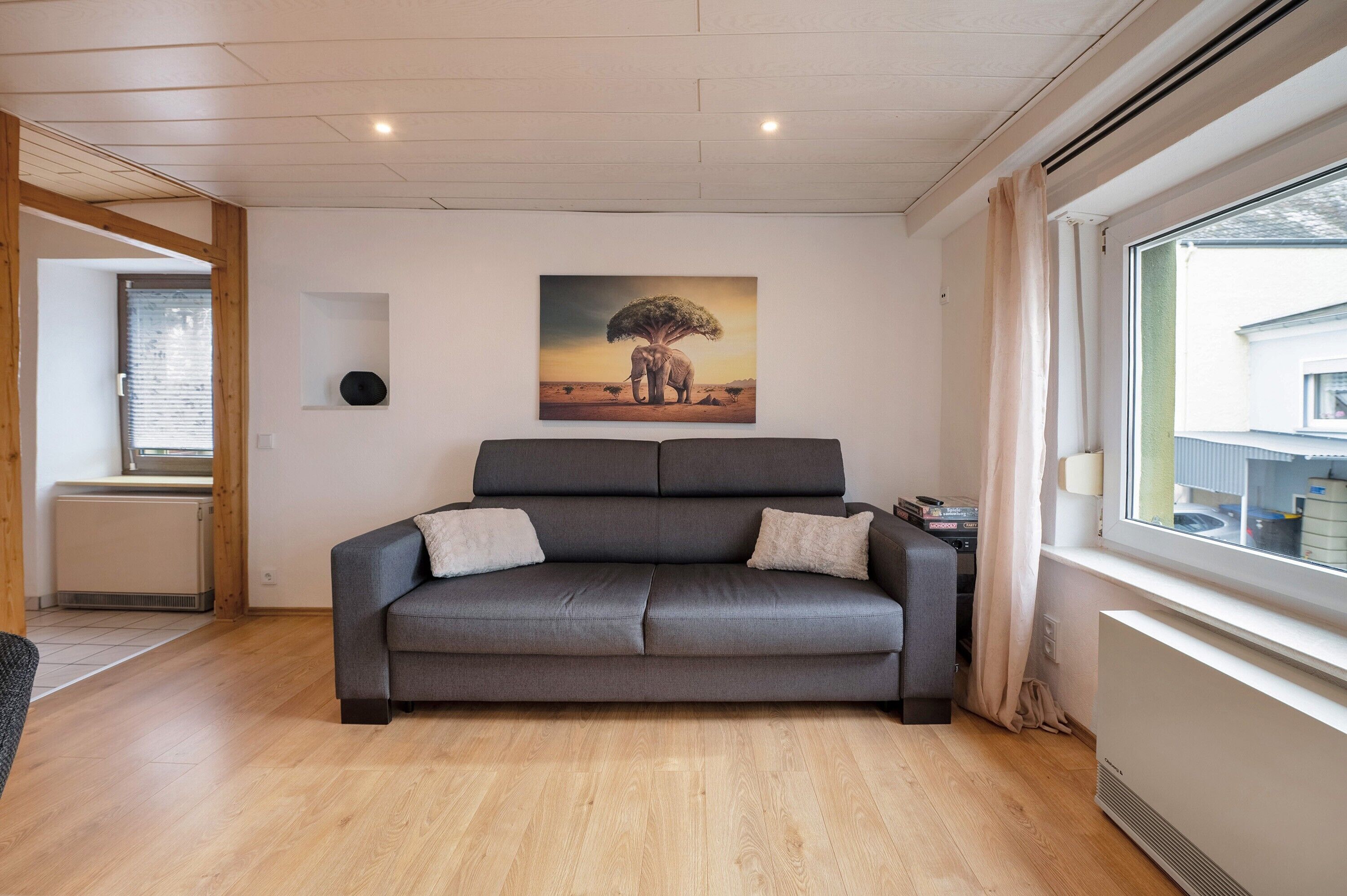 A modern living room with a grey sofa, a painting of an elephant, and a window with curtains.