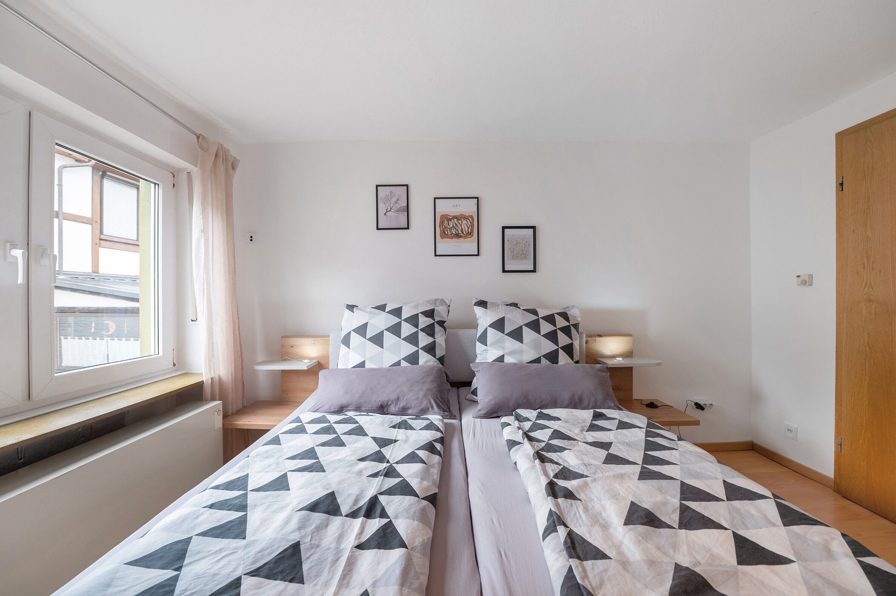 Une chambre avec deux lits, une fenêtre, une porte en bois et trois tableaux encadrés accrochés au mur.