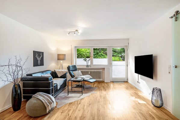 A modern living room with a sofa, armchair, and coffee table, a TV, and a window with blinds.