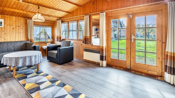 Een houten interieur met een bank, stoelen, een tafel en grote ramen.
