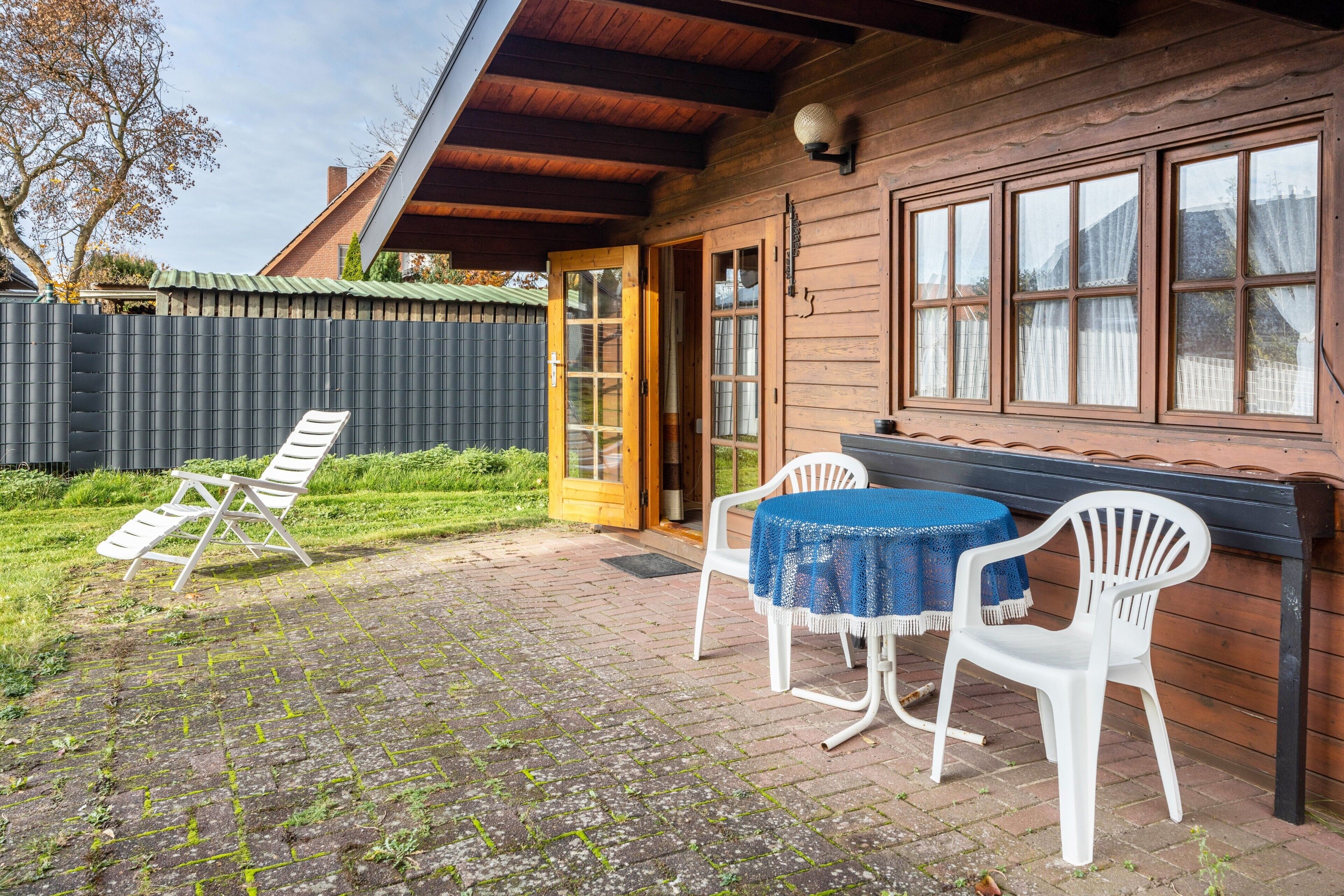 Une maison en bois avec un patio couvert, une table recouverte d’une nappe bleue et deux chaises blanches.