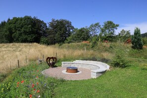 Área de estar ao ar livre circular com uma lareira central, rodeada por um muro de pedra e um campo gramado com flores silvestres.