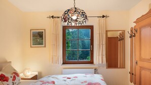Ein Schlafzimmer mit Bett, einem Fenster mit Vorhängen, einem Heizkörper, einem Nachttisch mit Lampe und einem Holzschrank.