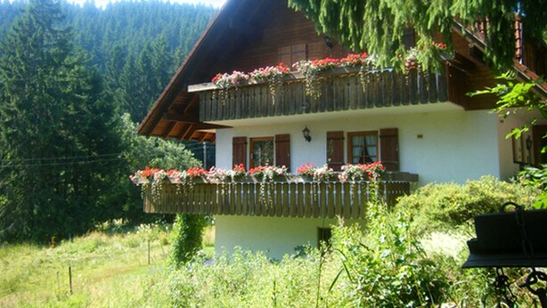 Ein traditionelles Chalet mit einem Balkon, der mit Blumen geschmückt ist, umgeben von üppiger Vegetation und einem bewaldeten Hintergrund.