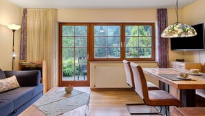 Ein Zimmer mit Esstisch, Stühlen, einer Couch, einem Fernseher und einem Fenster mit Blick auf Grünfläche.
