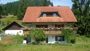 Ein zweistöckiges Haus mit rotem Ziegeldach, einem Balkon mit Blumen und einem bewaldeten Hang im Hintergrund.
