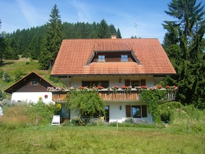 A two-story house with a red-tiled roof, a balcony with flowers, and a forested hillside in the background.