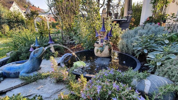 Ein Garten mit Teich, dekorativen Statuen und unterschiedlicher Bepflanzung.