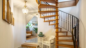 A spiral staircase with wooden steps and black railings, a white chair with a cushion, a patterned floor, and a chandelier. - Holiday Apartment 'Lavender' with Shared Garden, Balcony and Wi-Fi (Vogtsburg im Kaiserstuhl)