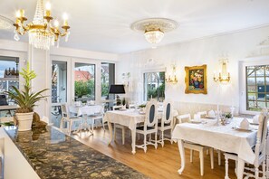 A dining area with white furniture, chandeliers, and a view through large windows.