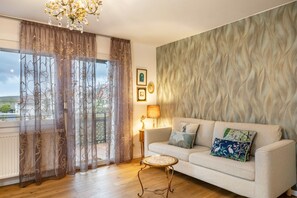 En stue med sofa, rundt bord, lampe og udsigt gennem lette gardiner.