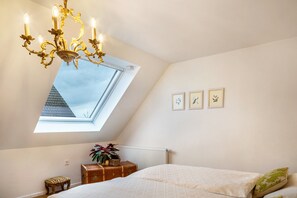 A bedroom with a skylight, a chandelier, a bed, a chest of drawers, and framed pictures on the wall.