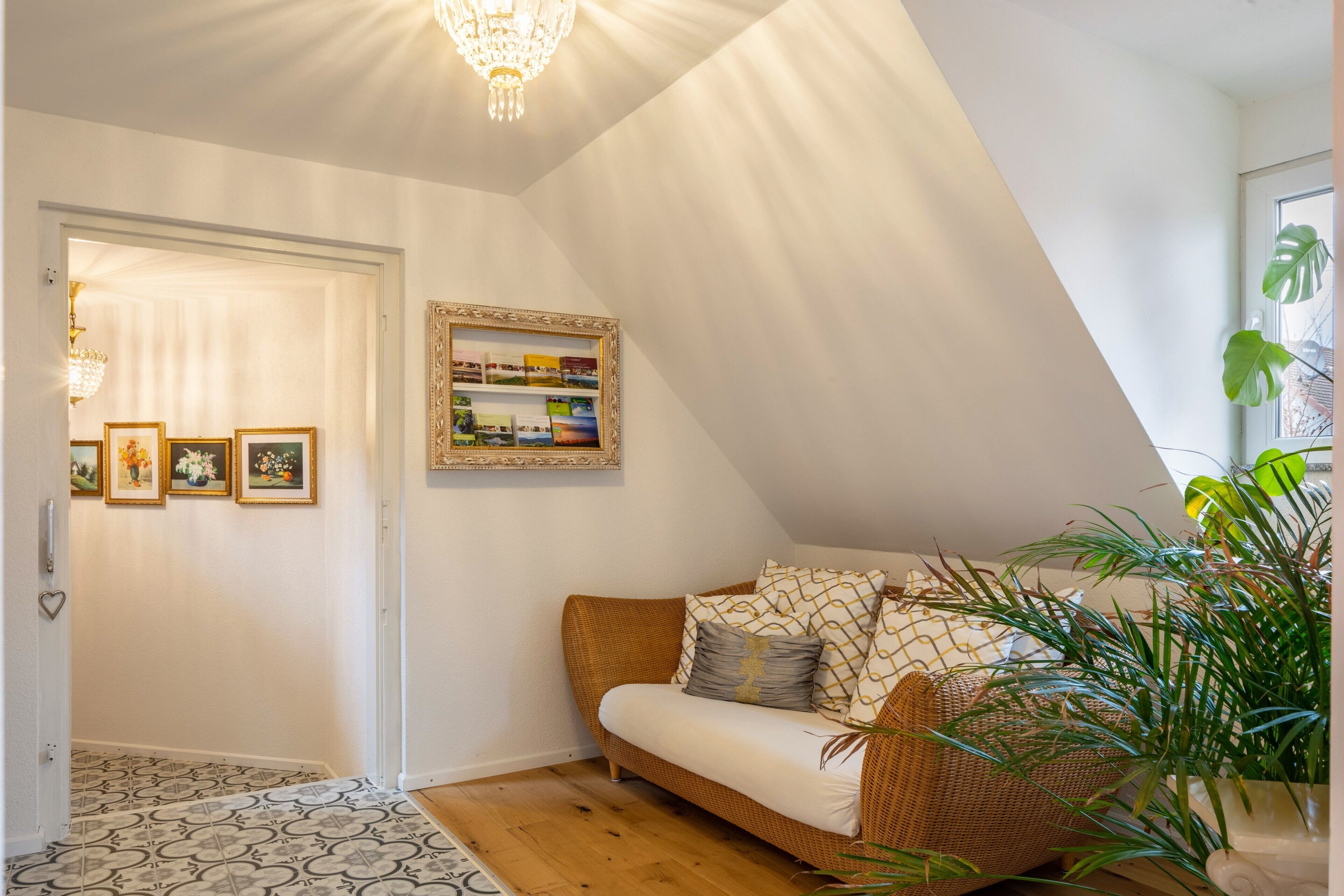 A cozy room with a sloped ceiling, a wicker sofa, a bookshelf, and a potted plant.