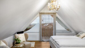 Een slaapkamer met een bed, een stoel, een klein tafeltje met een plant, een kroonluchter en uitzicht door een raam.