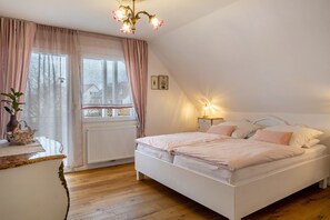 Ein Schlafzimmer mit Bett, Nachttisch, Kommode, Fenster mit Vorhängen und Heizkörper.