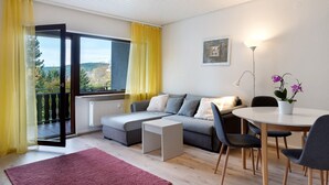 A modern living room with a sofa, a dining table, chairs, a small side table, a radiator, a window with curtains, and a view of the outdoors. - Apartment 'Bergblick' with Balcony and Wi-Fi (Triberg)