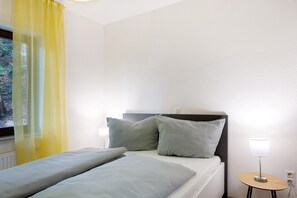 A hotel room with a bed, two pillows, a bedside table with a lamp, and a window with yellow curtains.