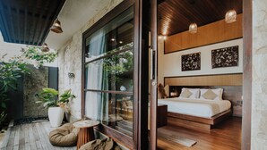 A modern bedroom with a large bed, wooden flooring, and a stone wall. There is a window with a view of greenery outside.