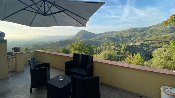 A balcony with a table and chairs, a large umbrella, and a scenic view of a valley and mountains. - Luxury Retreat in Montevettolini (Montevettolini)