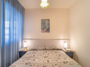 Un dormitori amb un llit, dues taules de nit, una làmpada de nit, un quadre a la paret i una finestra amb cortines blaves.