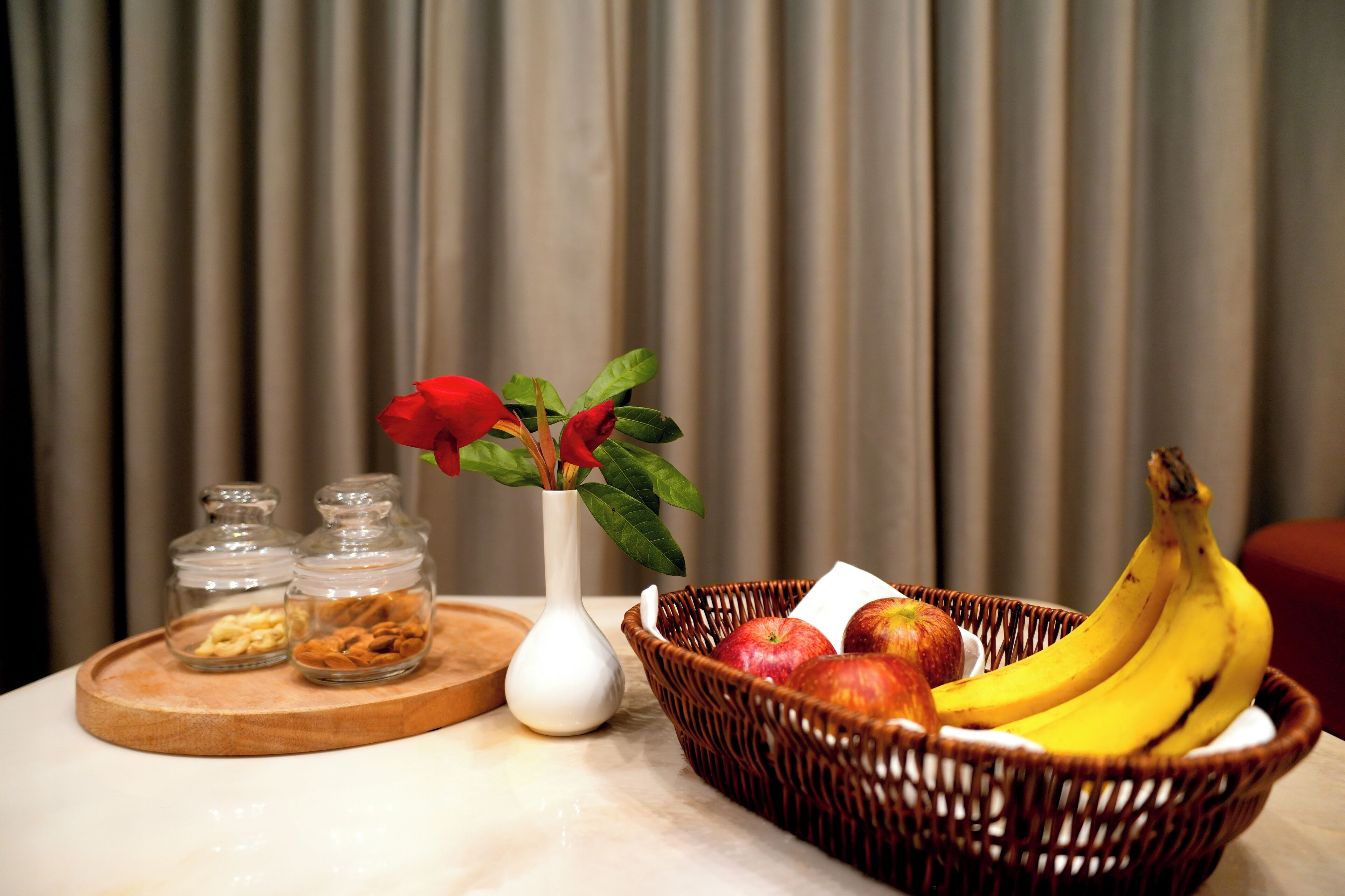 A table with a wicker basket containing bananas, apples, and a vase with red flowers.