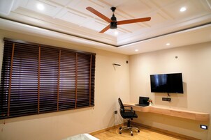 Una habitación moderna con ventilador de techo, un ventanal con persianas, un escritorio con computadora y un televisor montado.