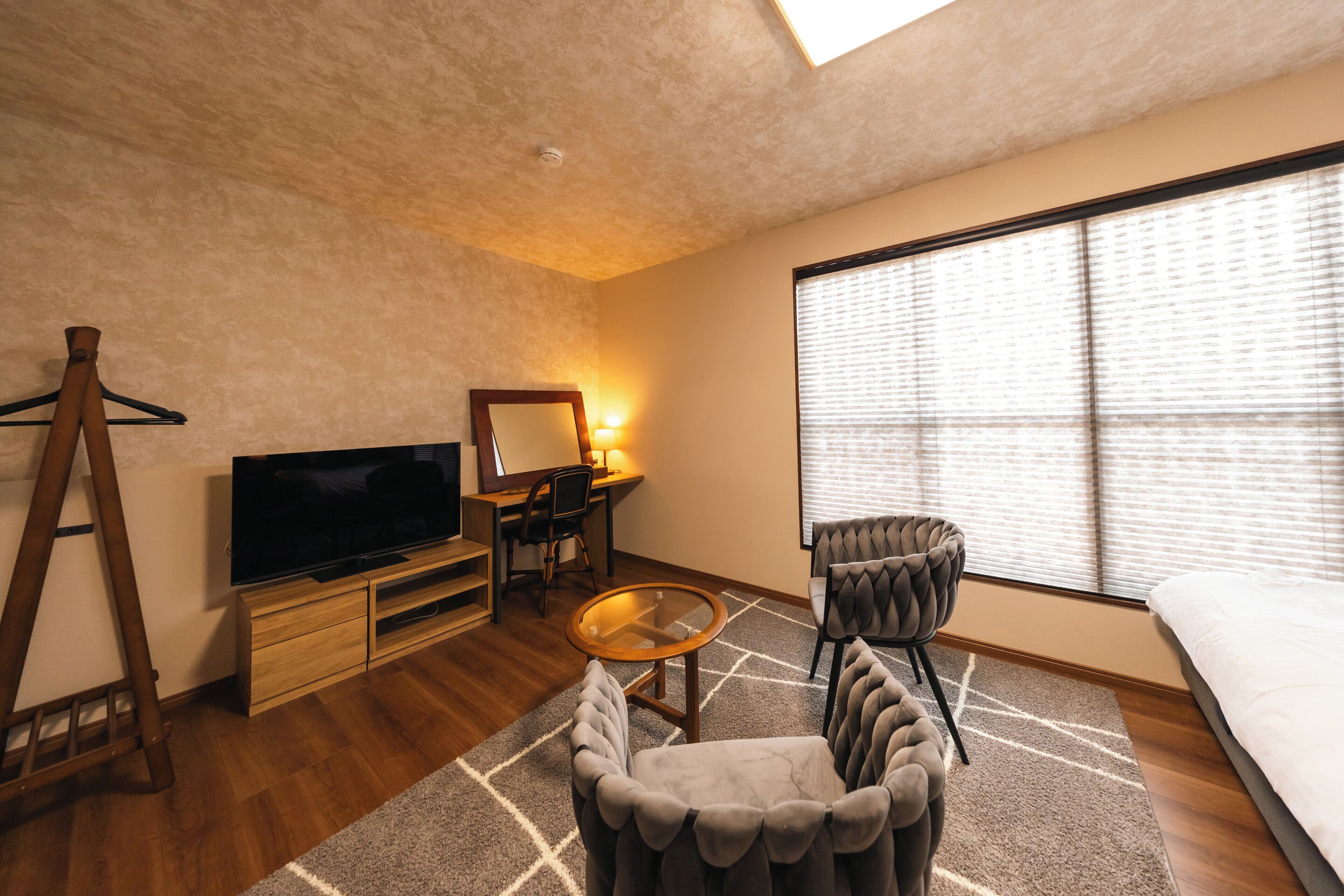 A modern room with a flat-screen TV, wooden furniture, a desk with a chair, a sofa, and a large window with blinds.