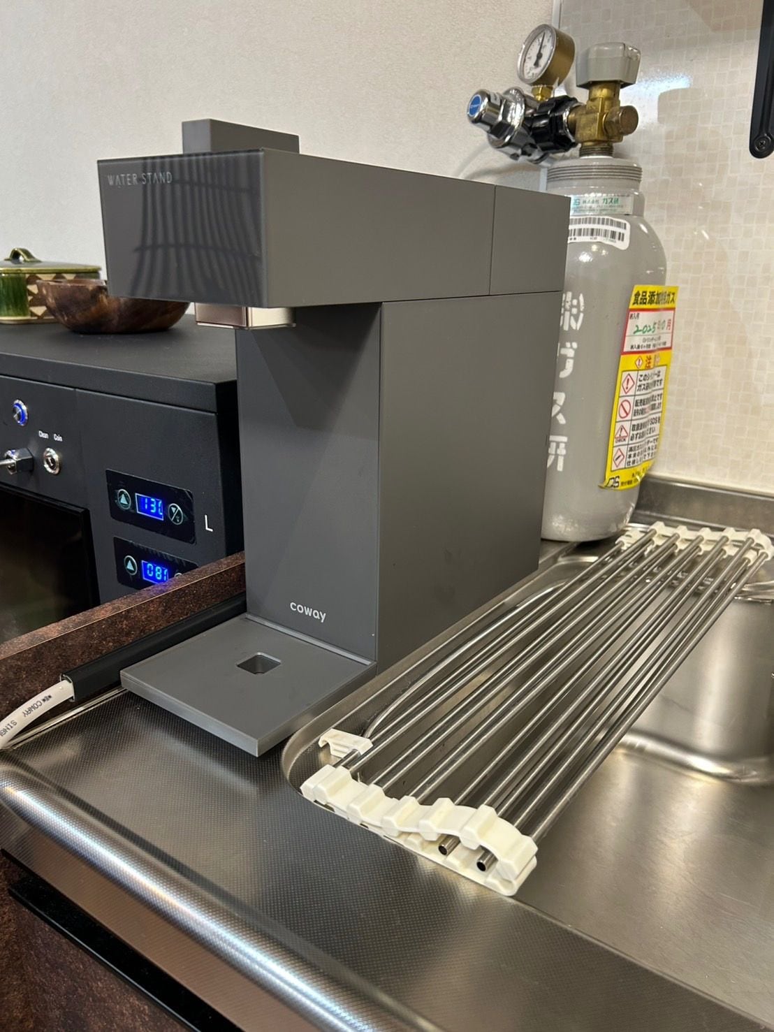 En kaffemaskine med digitalt display, en vanddispenser og en stålvask med et opvaskestativ.