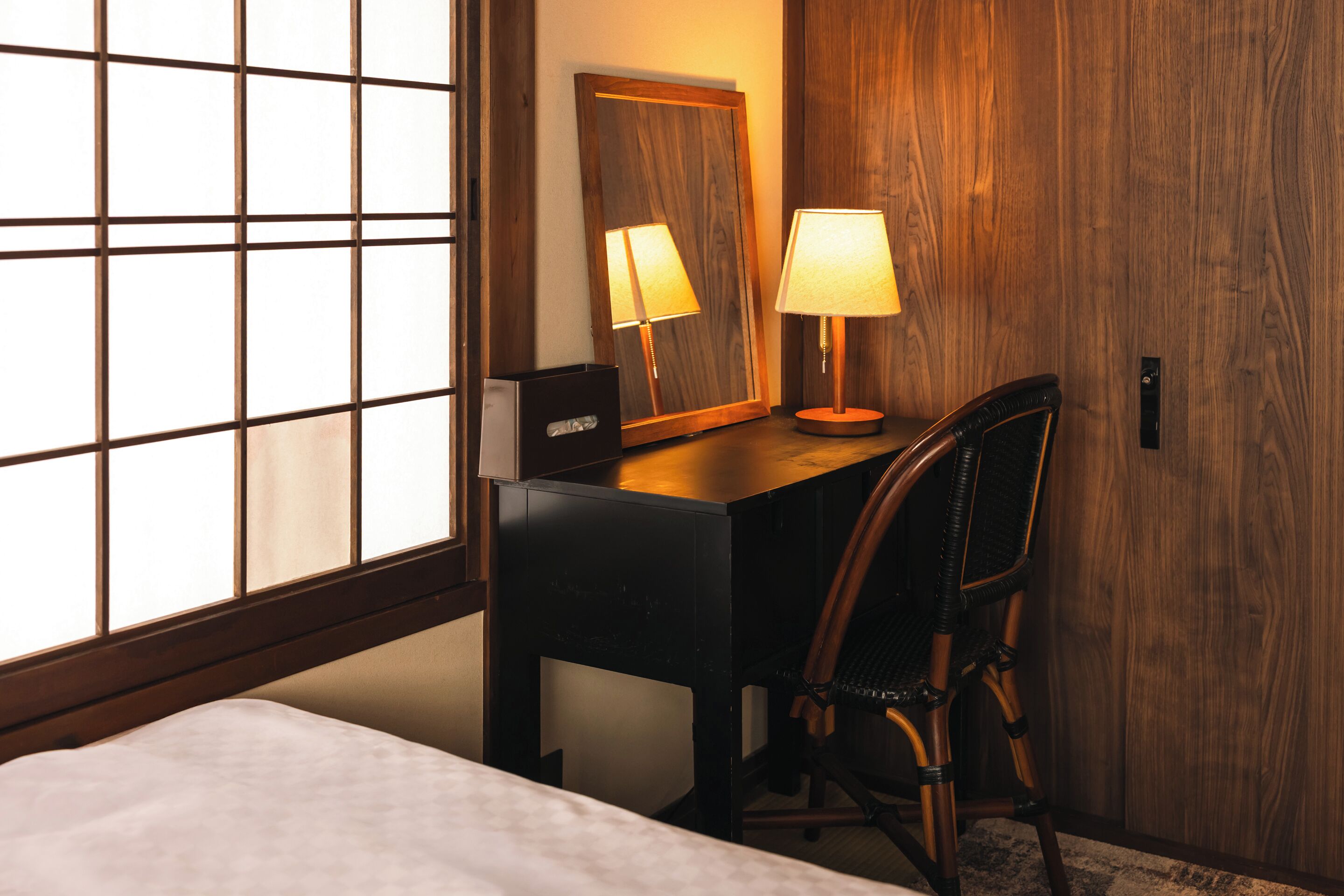 A hotel room with a bed, a desk with a chair, a lamp, a mirror, and a window with blinds.