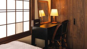 A hotel room with a bed, a desk with a chair, a lamp, a mirror, and a window with blinds.