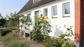 Una casa con fachada blanca, sección de ladrillo a la derecha y un jardín con girasoles.