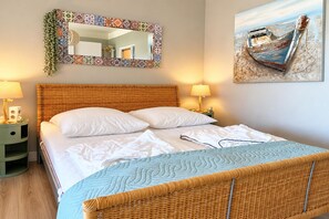 A bed with white linens, a wicker headboard, and a blue quilt. There is a mirror with a patterned frame and a painting of a boat on the wall.