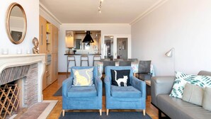 A modern living room with a fireplace, two blue armchairs with animal-themed cushions, a grey sofa, and a dining area in the background. - Apartment met zeezicht in Oostende (OOSTENDE)
